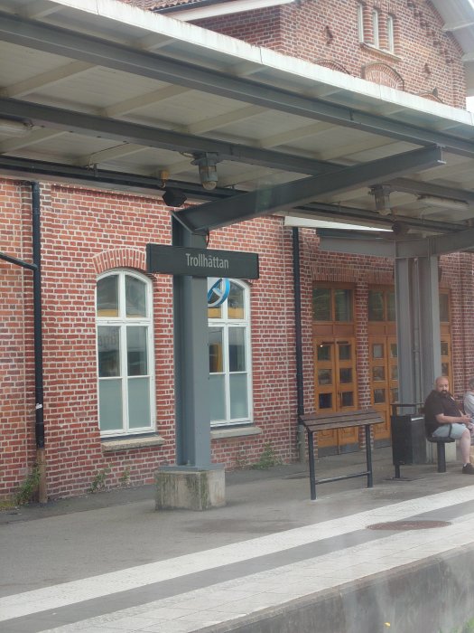 Skylt med texten "Trollhättan" vid järnvägsstation, tegelbyggnad och väntande person i bakgrunden.