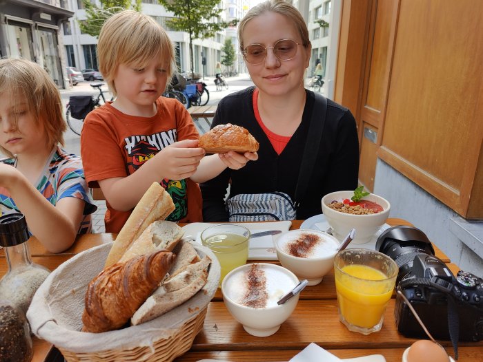 Familj som äter frukost utomhus med croissanter, baguette, yoghurt och juice.