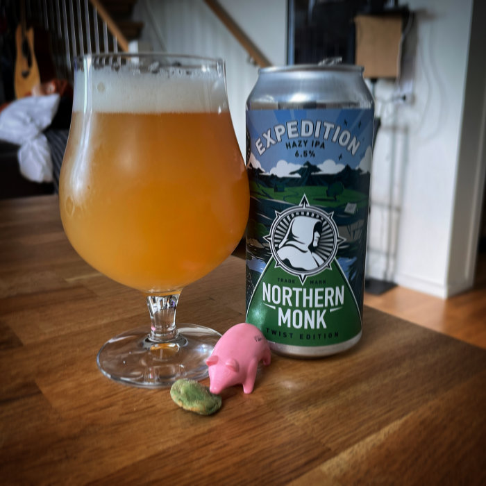 Ett glas med grumlig, guldfärgad öl intill en ölburk av märket 'Northern Monk' och en liten rosa plastgris på ett träbord.