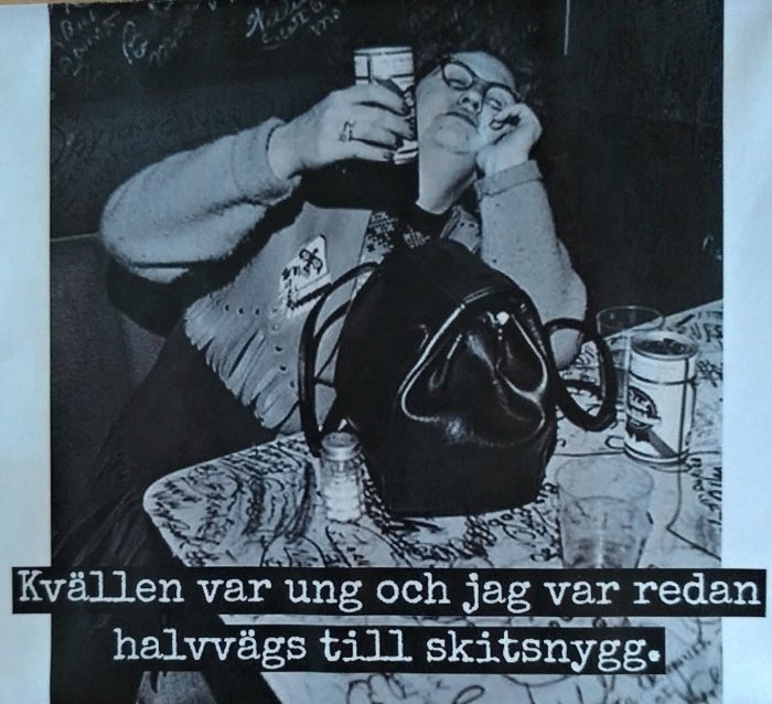 Kvinna med glasögon som håller ett glas nära sitt ansikte, sittandes vid ett bord med graffiti.