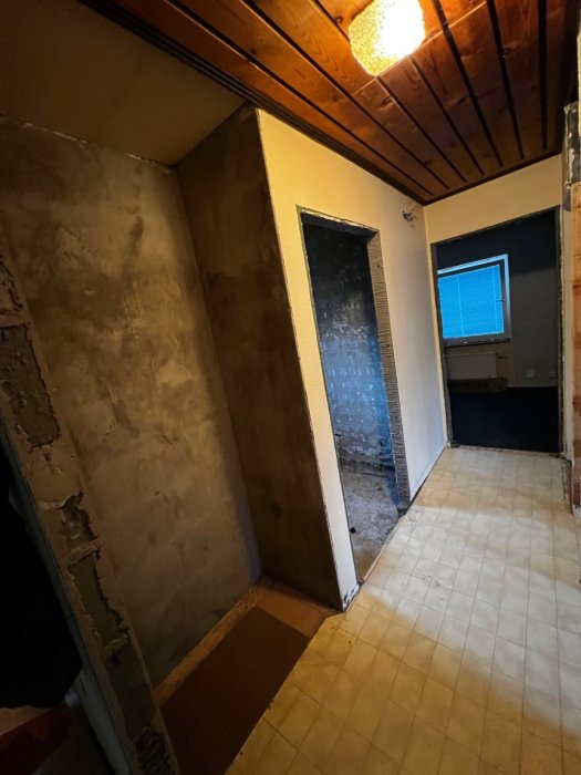 Renoveringsprojekt visar en korridor i ett hem med bar vägg efter rivning, trägolv och takbjälkar, med en öppen dörr.