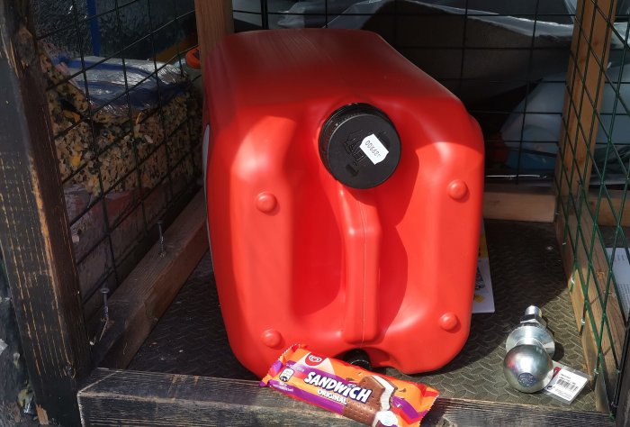 Röd bränsledunk på 25 liter från Aspen 2t, placerad i ett förråd med trädgårdsredskap.