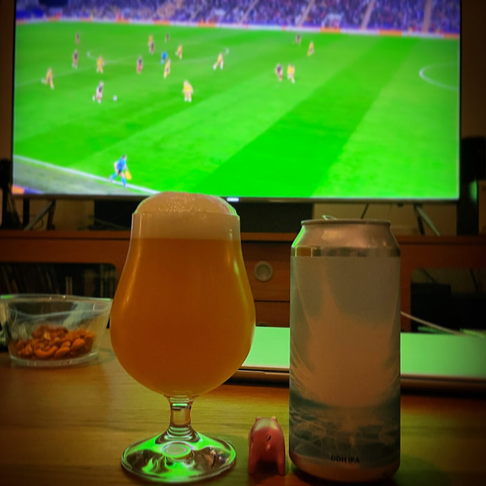 Ölglas och burk framför TV som visar en fotbollsmatch, med snacks och en liten rosa grisfigur i förgrunden.