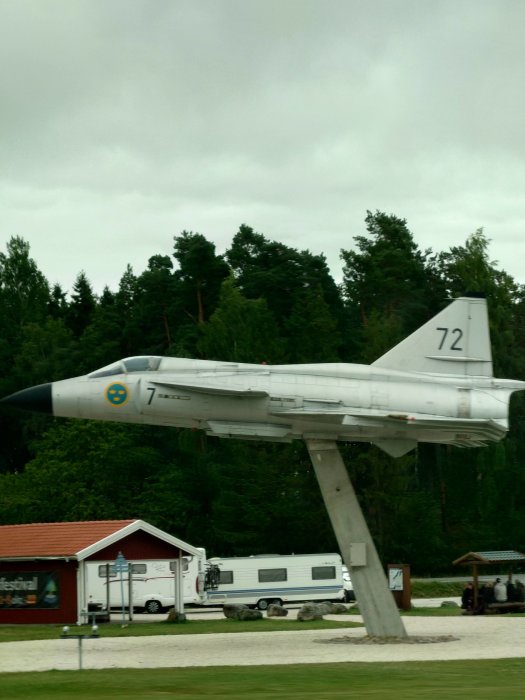 Ett uppmonterat stridsflygplan på en pelare utomhus med skog i bakgrunden och en husvagn vid sidan.