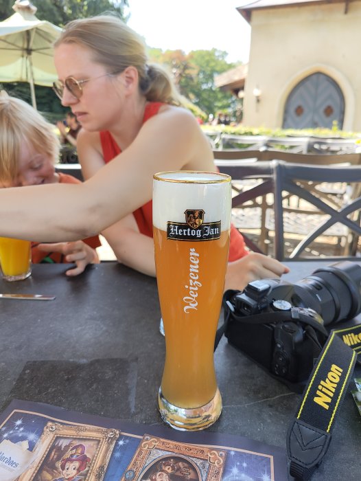 Ett glas veteöl i fokus med en kvinna och ett barn i bakgrunden, och en kamera på bordet.