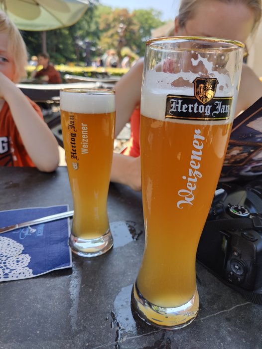 Två glas veteöl på ett bord med suddiga personer i bakgrunden.