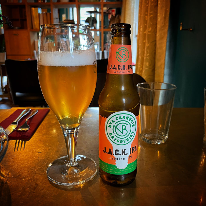 Ett glas öl och en flaska J.A.C.K. IPA på ett bord med en suddig bakgrund av en restauranginteriör.