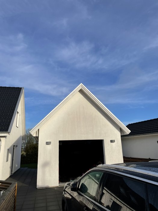 Vitputsad garagebyggnad med öppen svart port och del av bil under klarblå himmel.