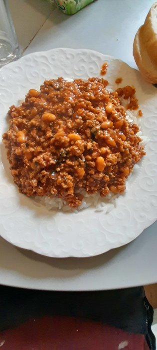 En tallrik med chili con carne över ris bredvid en bit bröd, på ett vitt fat.