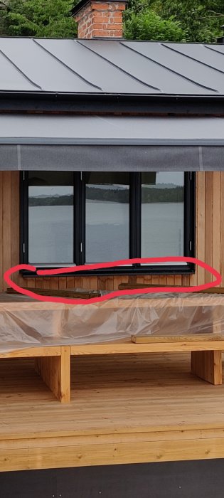 Utsikt från en tillbyggd veranda med stora fönster och ett trägolv, vatten och träd i bakgrunden, indikerat problemmarkerat med röd cirkel.