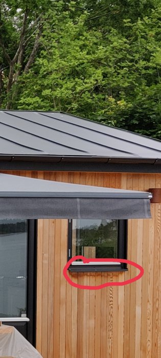 Parti av trävägg med fönster och ovanlig ventilationsspringa markerad med röd cirkel.