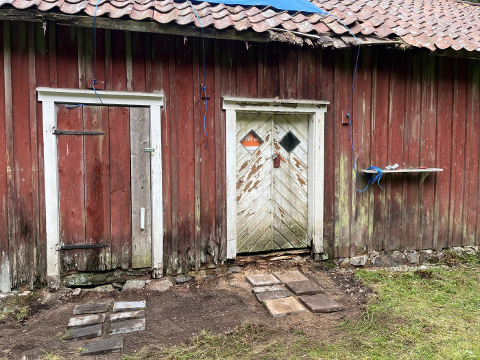 Rödmålad ladugård med två dörrar, den ruttna bron framför högra dörren utbytt mot brickade stenplattor.