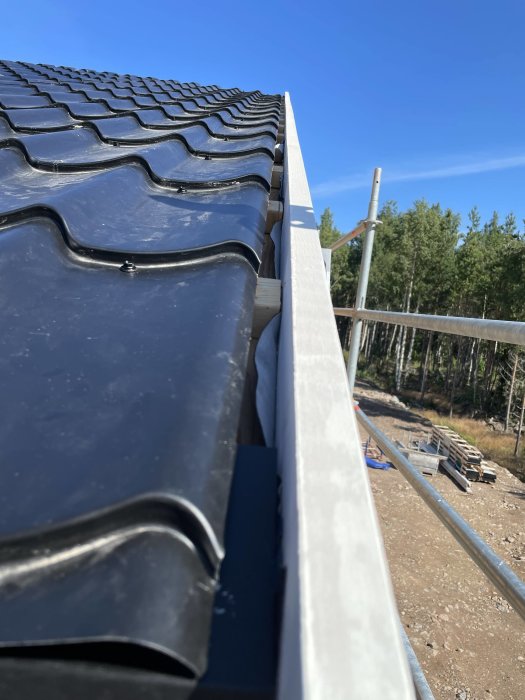 Svart takpannor längs med en vit takränna på en byggarbetsplats under en klarblå himmel.