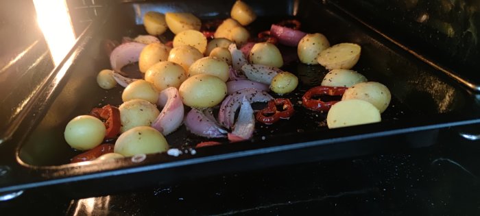 Färskpotatis, lök och tomat rostas i ugn på en svart plåt.