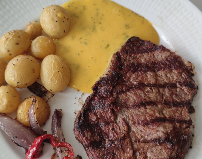 Grillad entrecote med rostad färskpotatis, lök och bearnaisesås på vit tallrik.