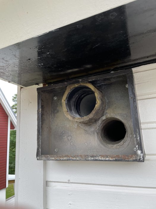 Bild på ett slitet svart ventilationssystem under takutsprånget på ett hus.