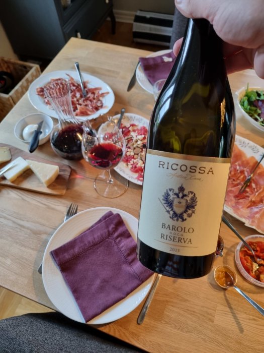 Hand håller flaskan Ricossa Barolo Riserva 2013 över dukat bord med charkuterier, ost, sallad och upphällda vinglas.