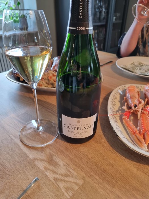 Flaska och glas med gyllengul champagne, etikett säger 2006 Castelnau blanc de blancs, mat i bakgrunden.
