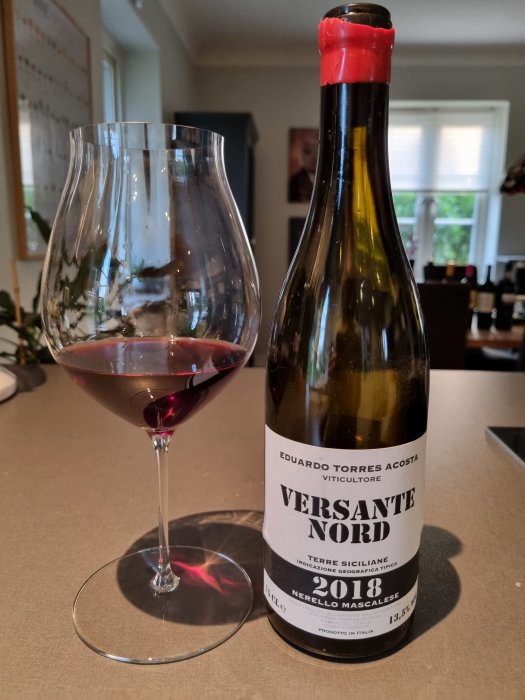 Flaska och glas med rödvin, etikett visar "Versante Nord 2018 Nerello Mascalese" på ett bord.