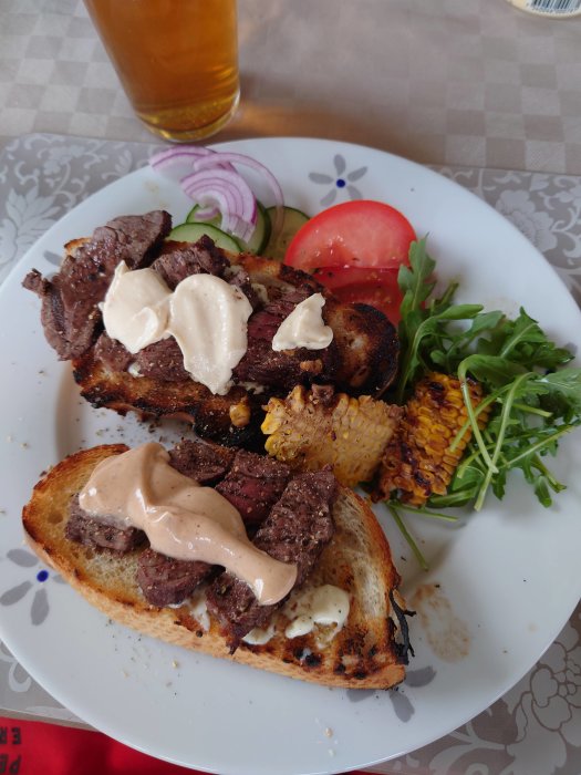 Öppen macka med skivat grillat kött och sås på grillat bröd, serverad med sallad och ölglas i bakgrunden.