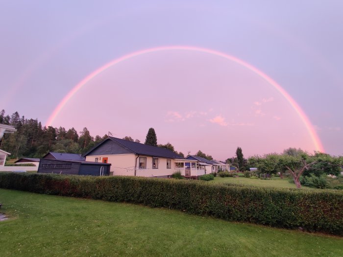 En dubbel regnbåge ovanför en vit villa med en trädgård vid skymningen.