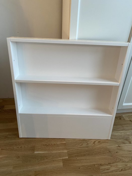 Vitt frittstående bokhylla med två hyllplan som står på ett trägolv mot en ljus vägg.