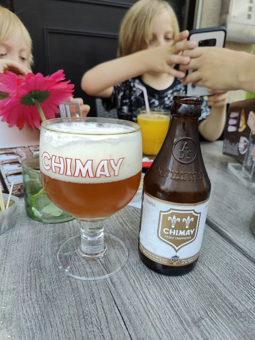 Chimay-öl i glas och flaska på bord med blomma, barn i bakgrunden dricker juice.