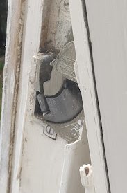 Närbild av ett fönsterlås på en vit fönsterram med trasigt fönsterglas.