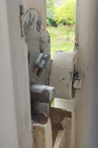 Utsikt genom ett gammalt fönster med flagnande färg och öppet hasp, trädgård i bakgrunden.