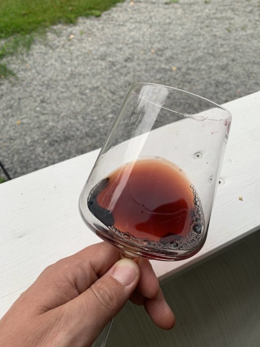 Ett vinglas hållet i handen fyllt med mörkt vin som står på en vit balkongkant, utomhusmiljö.
