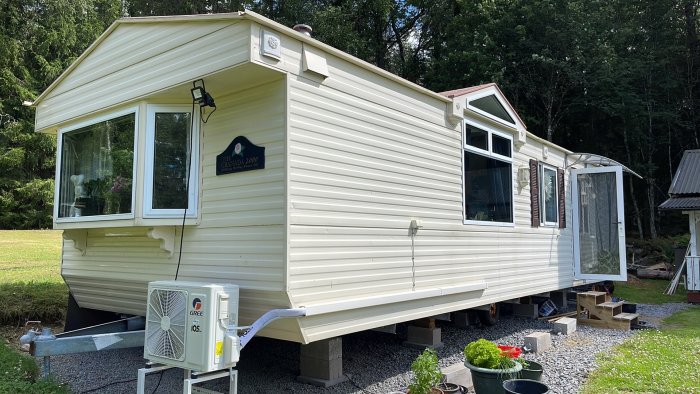 Husvagn stående på plintar omgiven av natur och med luftvärmepump framför.