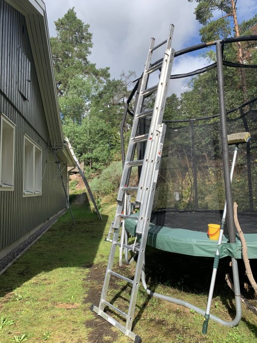 Aluminiumstege placerad mot husvägg bredvid studsmatta, med mopp och hink på gräsmatta.