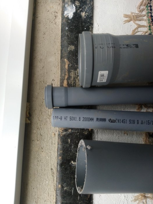 Grå avloppsrör i PVC ligger på en smutsig träbräda vid en vit vägg.