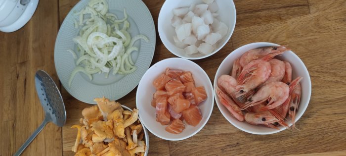 Ingredienser till fiskgryta: skivad lök, kantareller, rå lax och scampi i skålar på ett träbord.