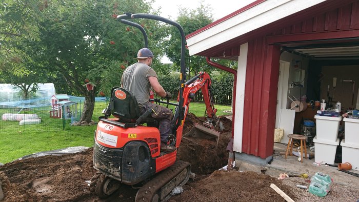 Person använder minigrävare för att gräva för avlopps- och elinstallationer nära ett garage.