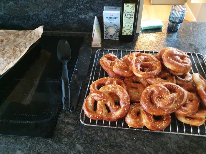 Hembakade pretzels på en kylgaller bredvid olivolja och flingsalt på ett köksbänk.