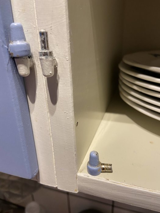 Avbruten skruv med platt yta på änden ligger bredvid ett skåps gångjärn på vitmålad yta, med tallrikar i bakgrunden.