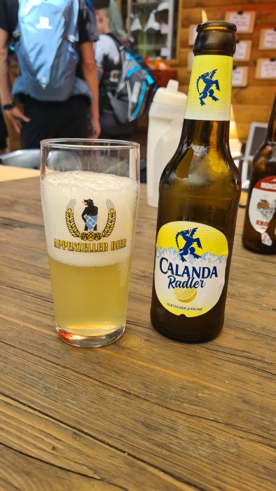 Ett glas öl och en flaska Calanda Radler på ett träbord med vandrare i bakgrunden.