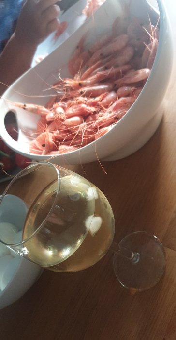 Skål med räkor och ett glas vitt vin på träbord, med en person i bakgrunden.