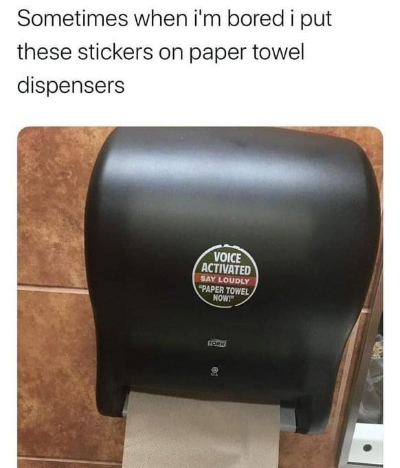 Svart pappershandduksautomat med skämtsamt klistermärke "Voice Activated. Say Loudly 'Paper Towel Now!'
