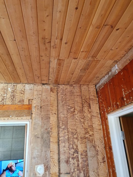 Träpanel på vägg och tak i olika skeden av renovering, med synlig isolering och dörröppning.