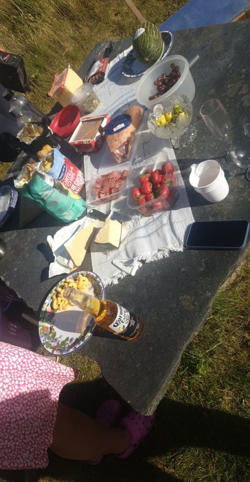 Sommarpicknick med ost, kex, melon, jordgubbar och dryck på en stenplatta utomhus.