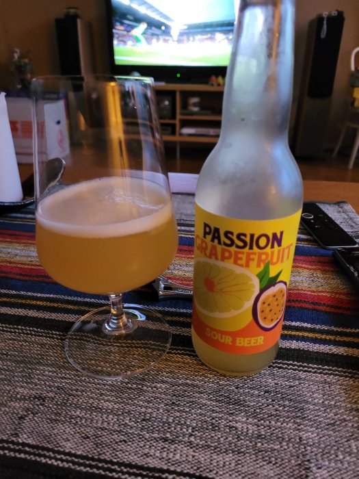 Ölglas och flaska med etiketten "Passion Grapefruit Sour Beer", framför en TV som visar sport.