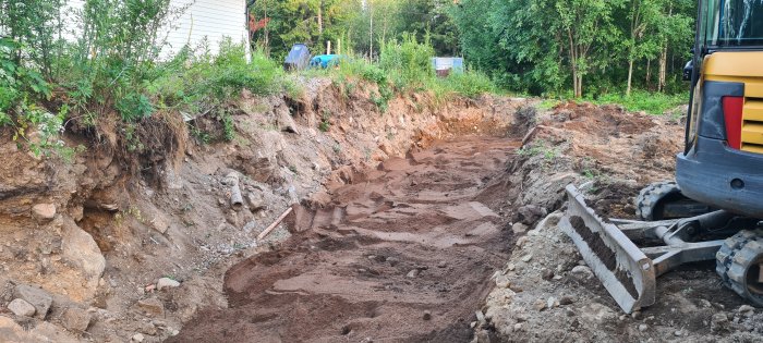 Grävmaskin vid öppnad dräneringsgräv med utspridd dränsand och synlig fördelningsbrunn i bakgrunden.