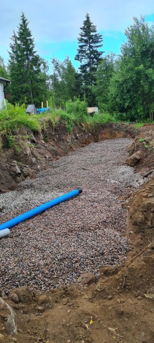 Grusfylld fördjupning med blå fördelningsrör och synlig dränsand för grundförstärkning i ett byggprojekt.