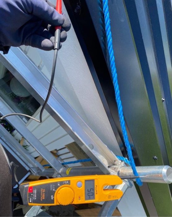 Hand håller en skruvmejsel nära en metallprofil med en multimetrar som mäter ström vid sidan.