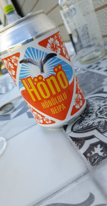 Burk med öl märkt "Hönö Honolulu NEIPA" på ett bord med mönstrad duk.