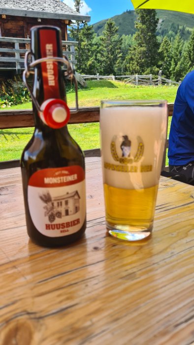 Flaska av Monsteiner Huusbier bredvid ett glas Weissbier på ett träbord utomhus.