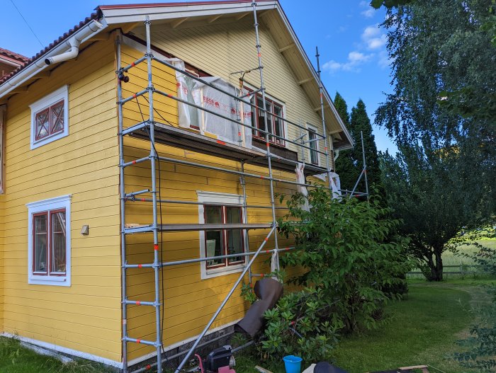 Byggställning mot gult hus under renovering med påbörjad fasadpanel och vindskydd.