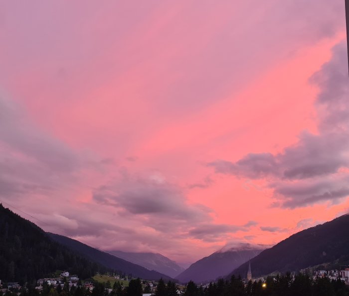 Dramatisk rosa och lila himmel vid solnedgång över ett bergslandskap med silhuetter av träd och byggnader.